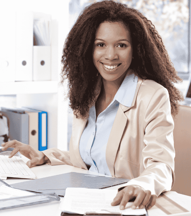 A smiling professional seated at a desk in a bright office, surrounded by documents and organised workspace items.