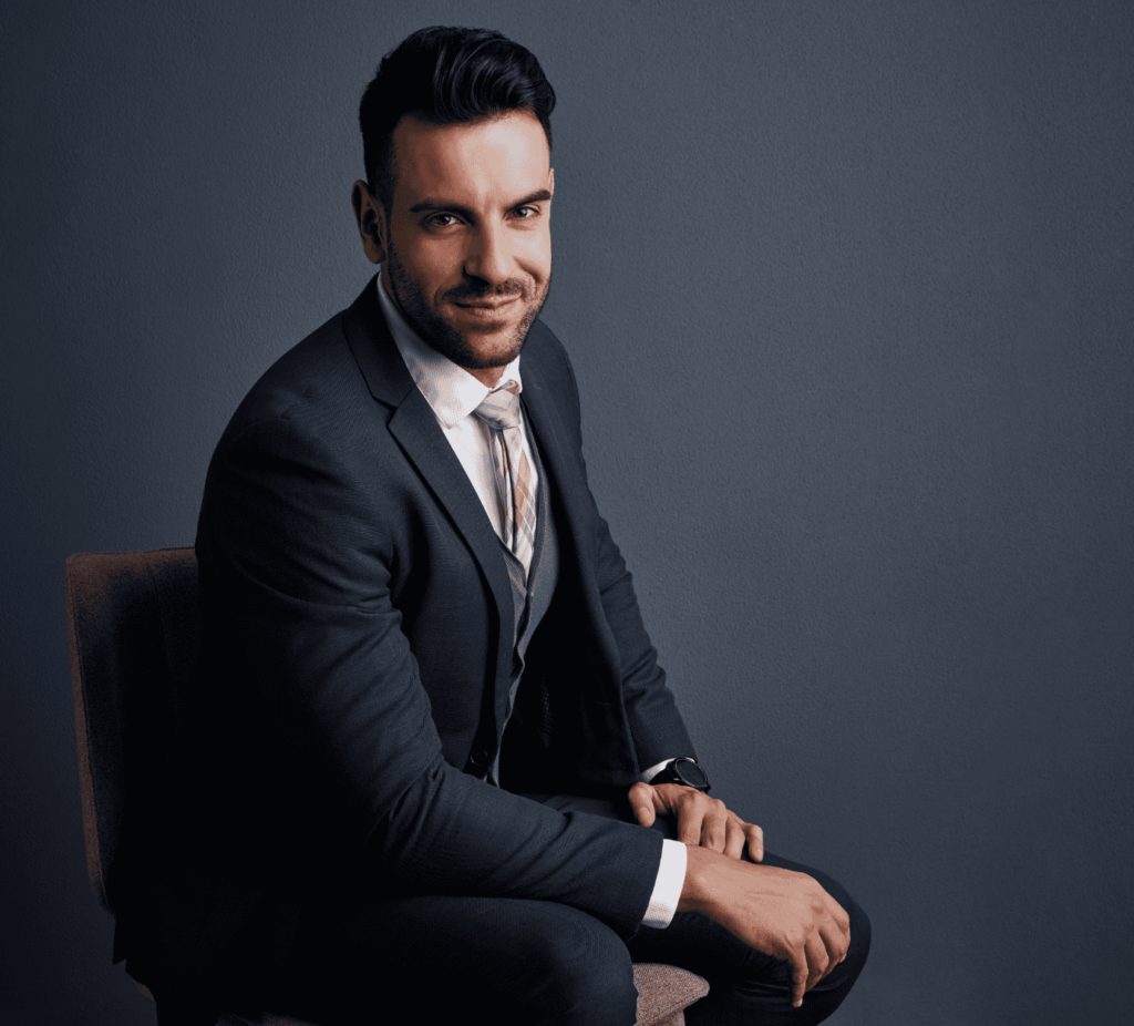 A professional seated against a dark background, wearing a formal suit and looking confidently at the camera