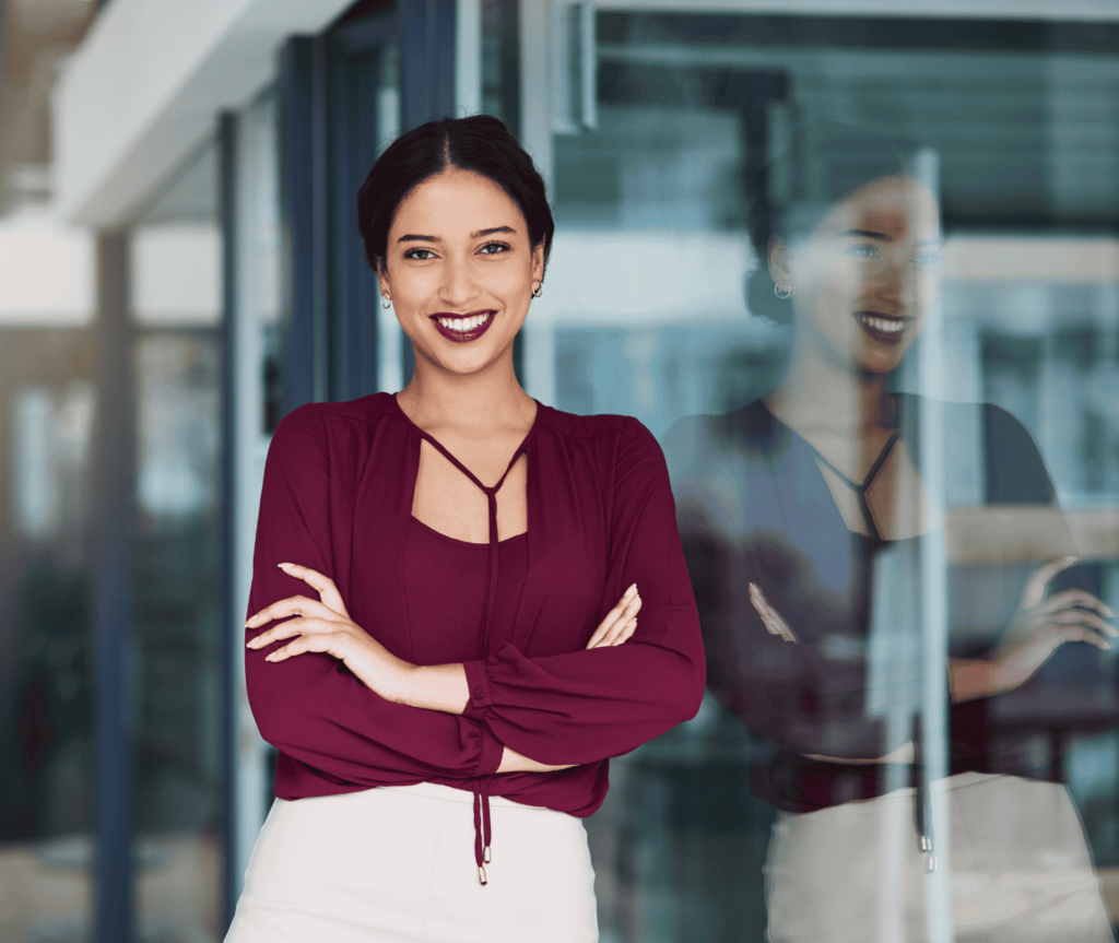 A confident professional standing indoors with arms crossed, wearing a burgundy blouse and white trousers, conveying assurance and clarity.