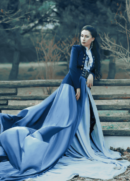 A person in an elegant blue gown and tailored jacket leaning on a wooden fence in a forest setting, conveying sophistication and exclusivity.